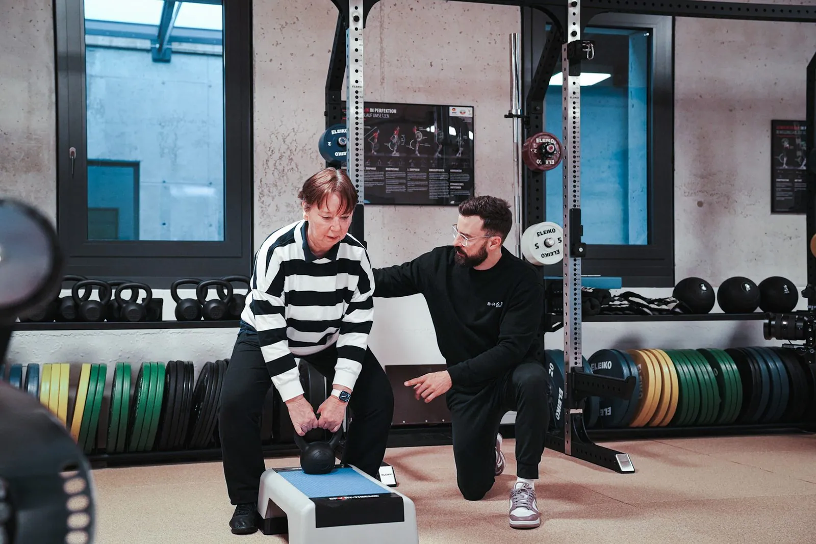 Trainer leitet eine Seniorin beim Kettlebell-Heben auf der Trainingsfläche im BRIKS Studio an